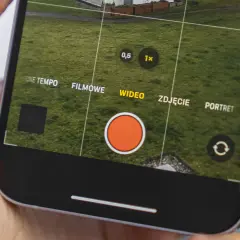 Dłoń trzyma iPhone'a z otwartą aplikacją aparatu, widoczny przycisk nagrywania wideo.