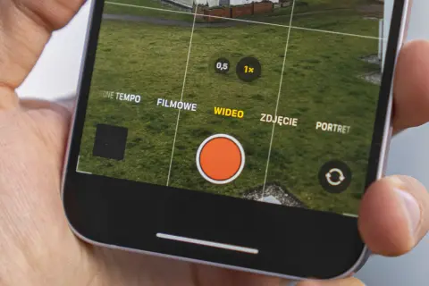 Dłoń trzyma iPhone'a z otwartą aplikacją aparatu, widoczny przycisk nagrywania wideo.